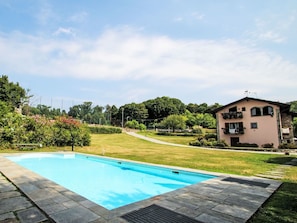 A swimming pool with a stone border, surrounded by a grassy area and a building with a balcony. - Natalina by Interhome (Cocquio Trevisago)