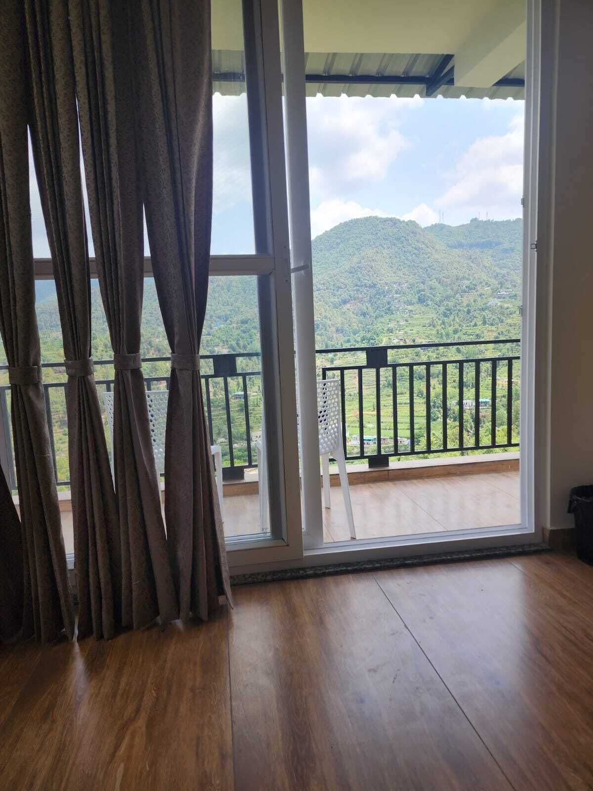 Una habitación con piso de madera, una puerta corrediza de vidrio que da a un balcón con baranda de metal y vistas a un paisaje verde con colinas.