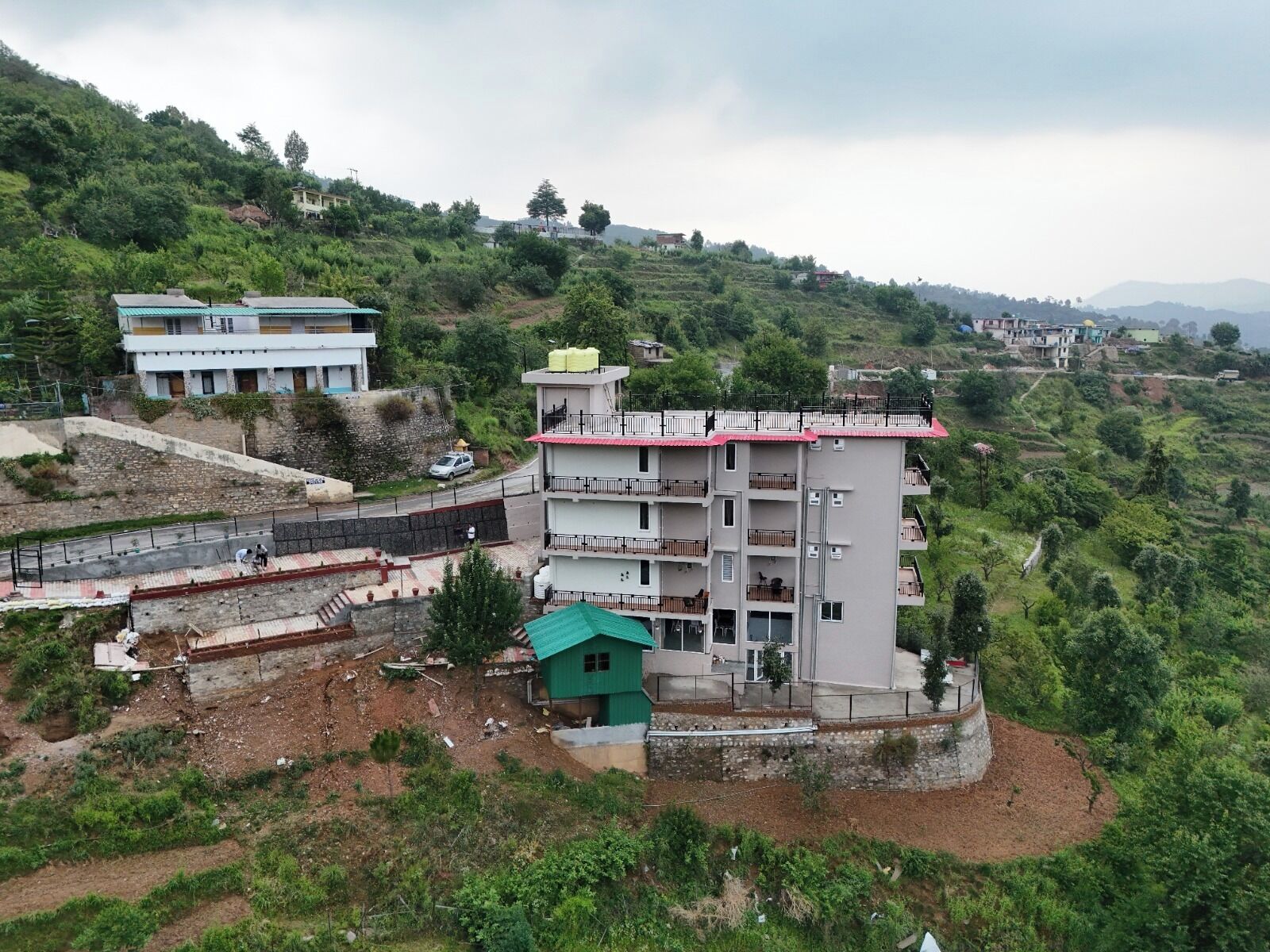 Un edificio de varios pisos con balcones, ubicado en una ladera con una carretera y vegetación al fondo.