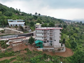 A multi-story building with balconies, situated on a hillside with a road and greenery in the background. - Mountain Stories Mukteshwar (Dhari)