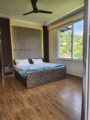 Quarto com cama grande, ventilador de teto, mesa de cabeceira e janela com vista para o verde.