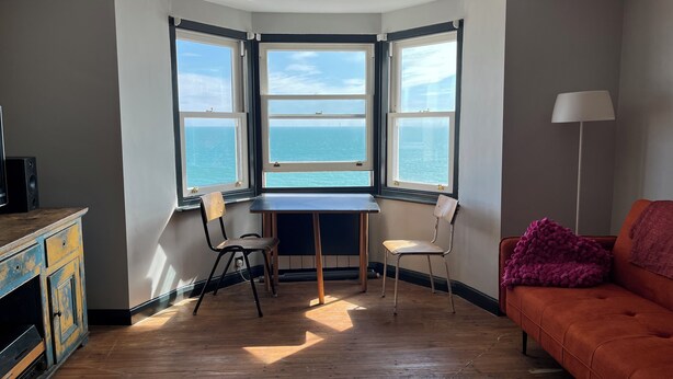 Ett rum med ett hörnskrivbord, två stolar, en soffa och utsikt över havet.