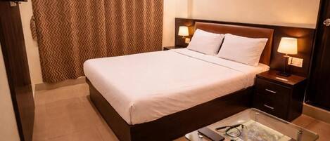 Una habitación de hotel con una cama, mesitas de noche, una mesa de centro de vidrio y una cortina.