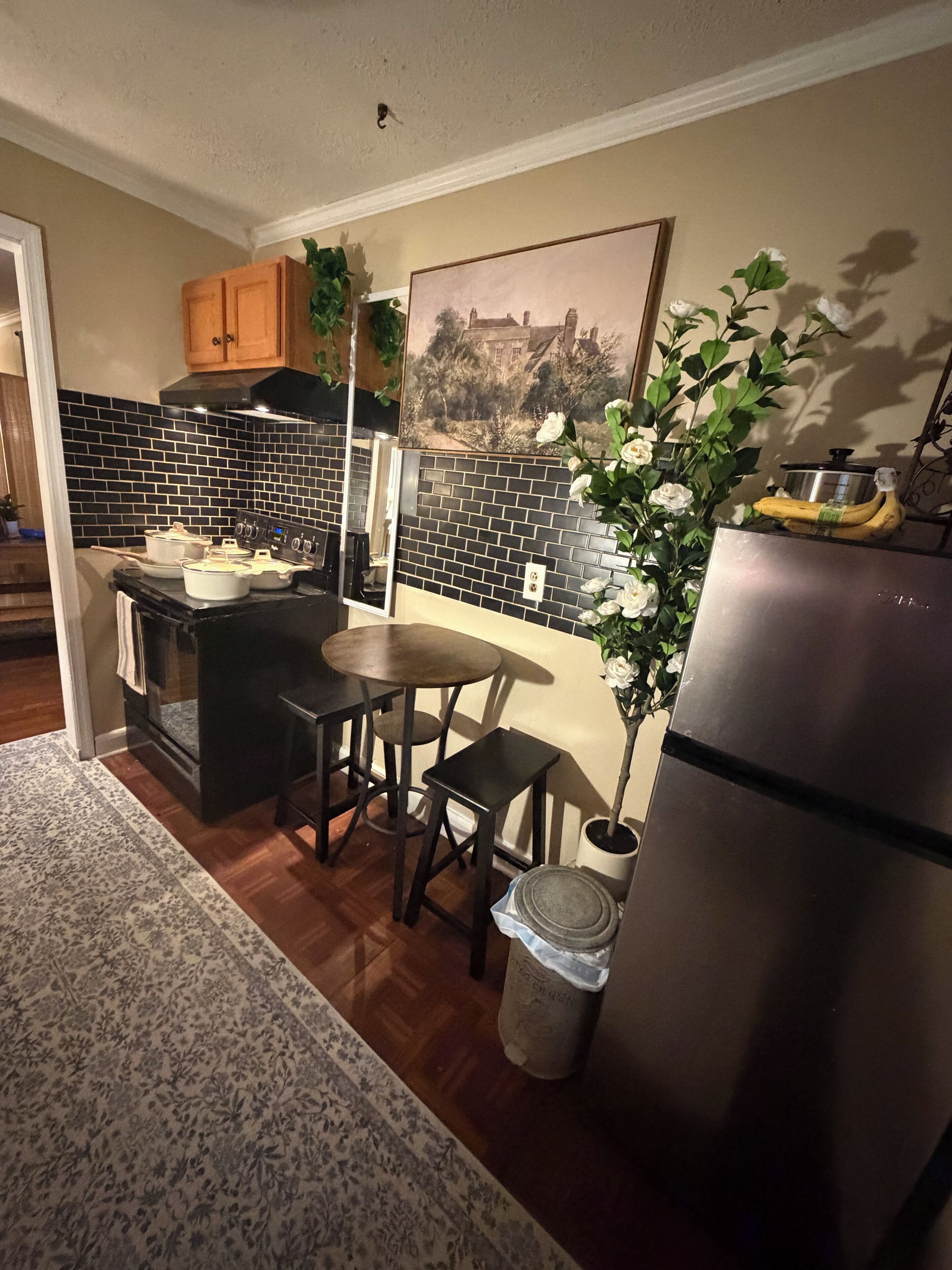 A compact kitchen with a stove, sink, and refrigerator. There is a dining table and chairs, a trash bin, and a potted plant.