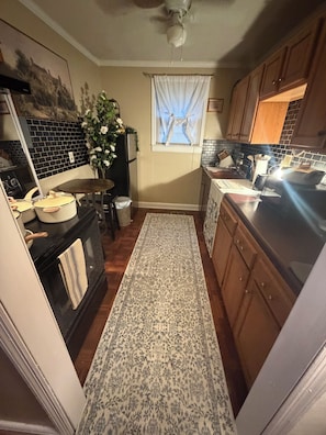 A kitchen with a black countertop, wooden cabinets, a patterned rug, and a window with curtains.