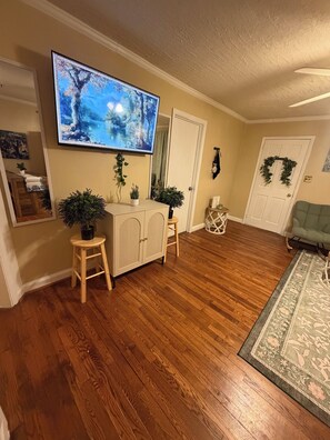 A living room with a wooden floor, a television mounted on the wall, a green armchair, and a decorative rug. - Gorgeous 3-bedroom bungalow with Hot Tub, Games and AC in Southern Pines (Southern Pines)