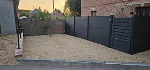 A gated backyard with gravel, a wooden fence, and a brick building in the background. - Le Petit Nid: Private spa & movie theater—a romantic and unforgettable experience for two (Courtonne-les-Deux-Églises)