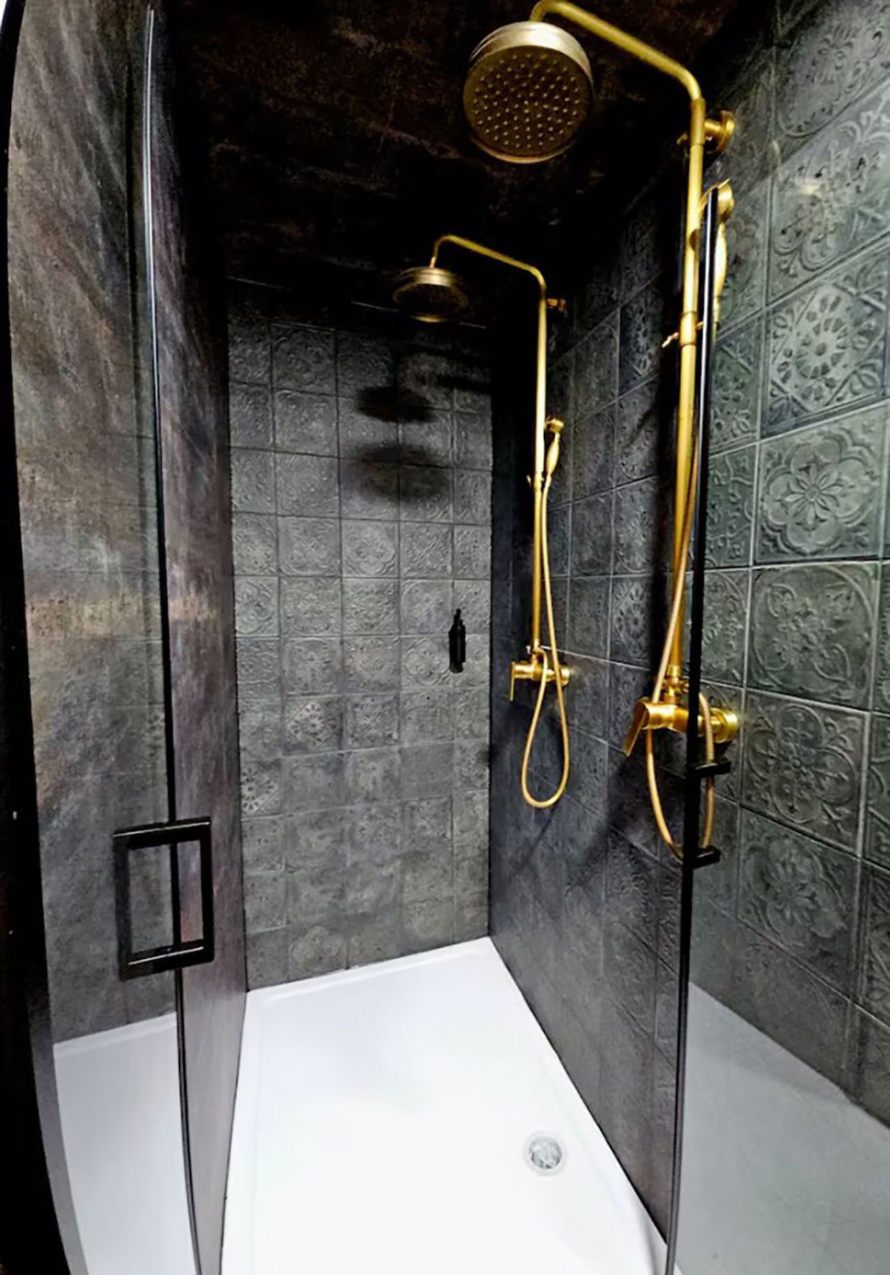 Eine Dusche mit goldfarbener Duschbrause und Duschschlauch, vor dunklen, gemusterten Fliesen.
