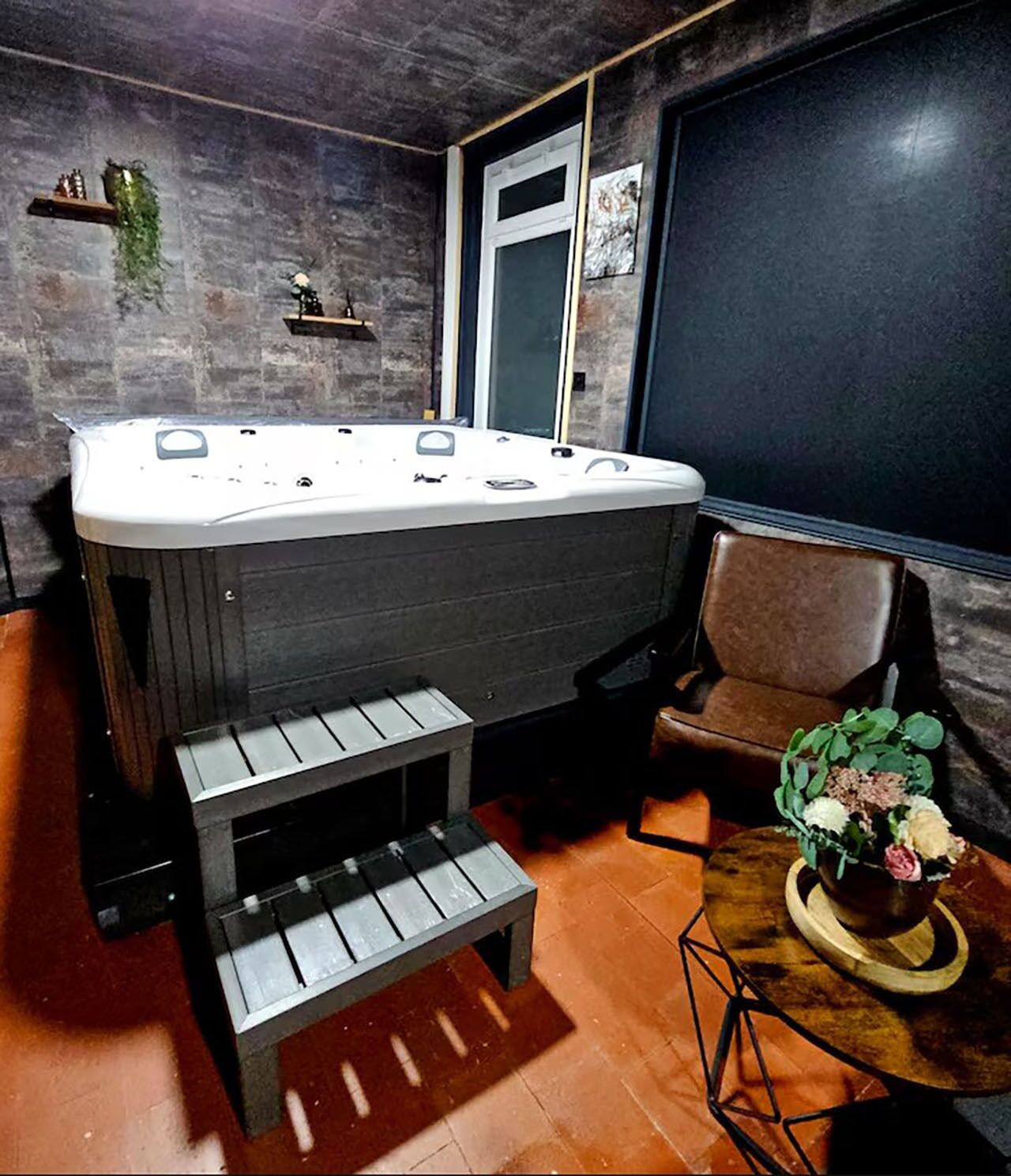 A room with a hot tub, a chair, a small table with a plant, and a window with a view.