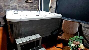 A room with a hot tub, a chair, a small table with a plant, and a window with a view.