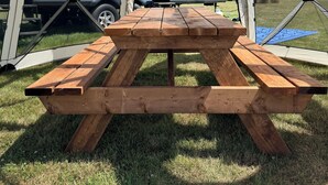Une table de pique-nique en bois, placée sous une structure en forme de tente, avec une vue sur une personne installée dans une tente à l’arrière-plan.