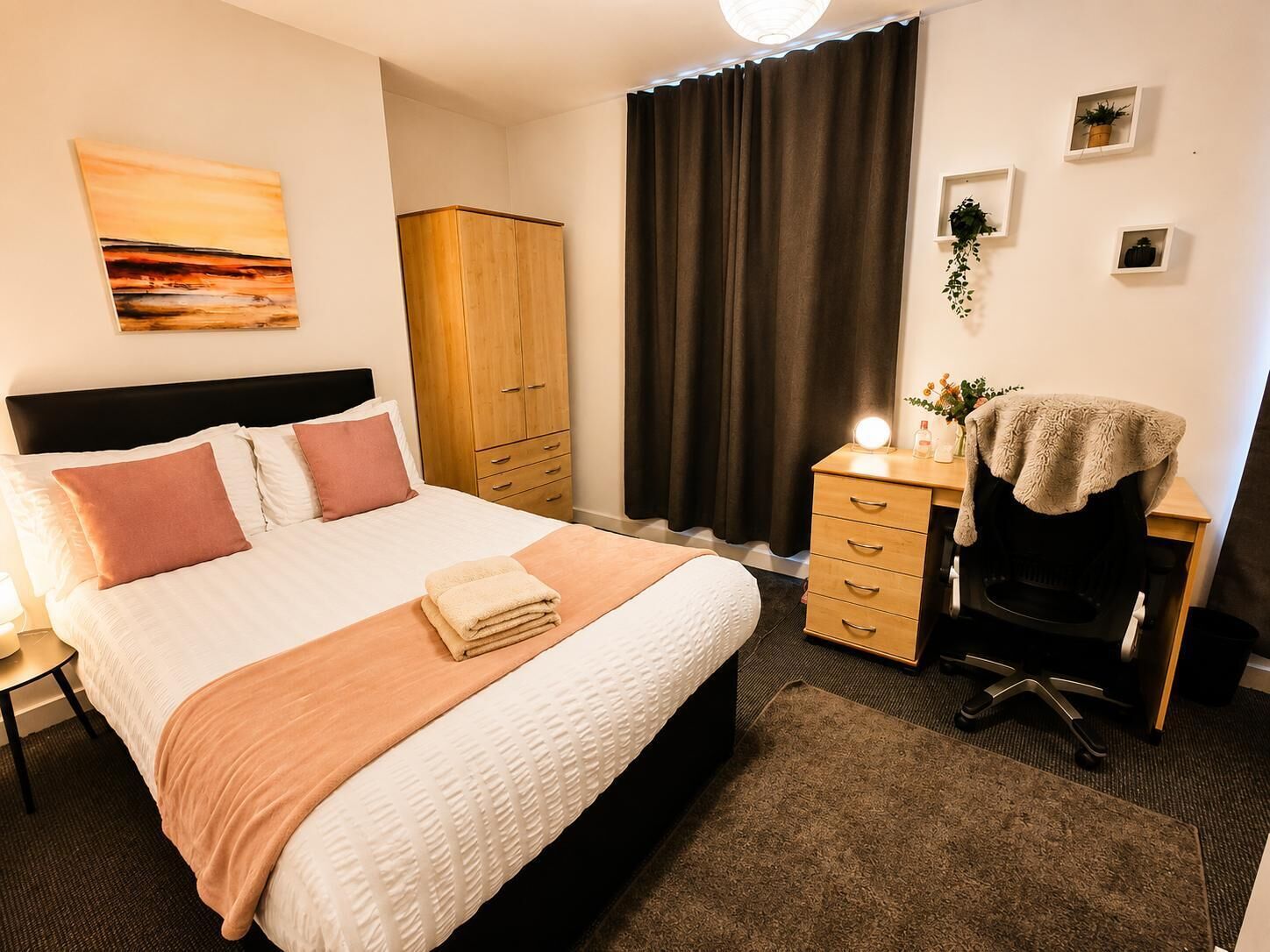 A hotel room with a bed, a wardrobe, a desk with a chair, a desk lamp, and a wall-mounted light.