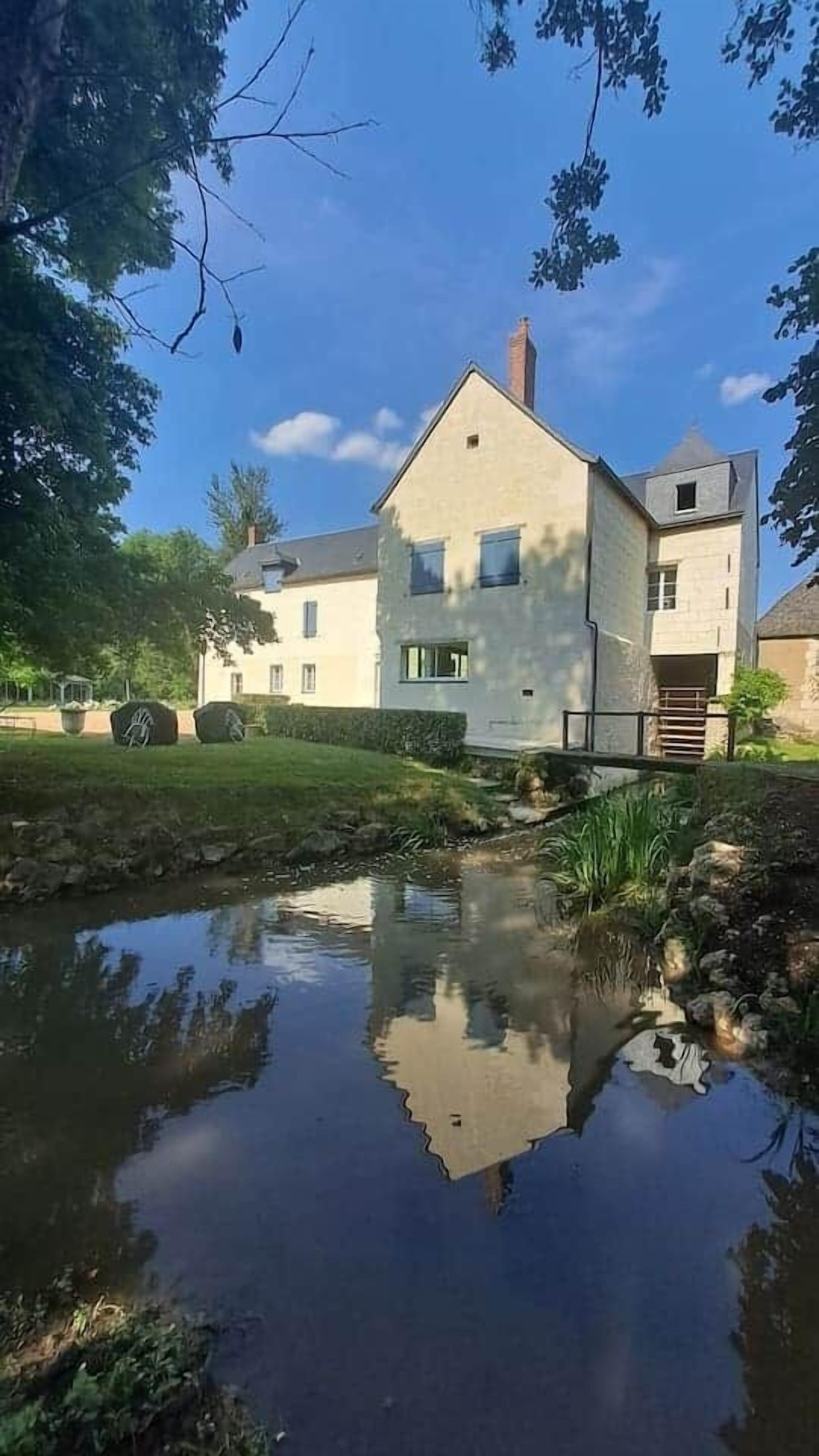 Une maison à deux étages avec un étang devant, entourée d’arbres et sous un ciel dégagé.