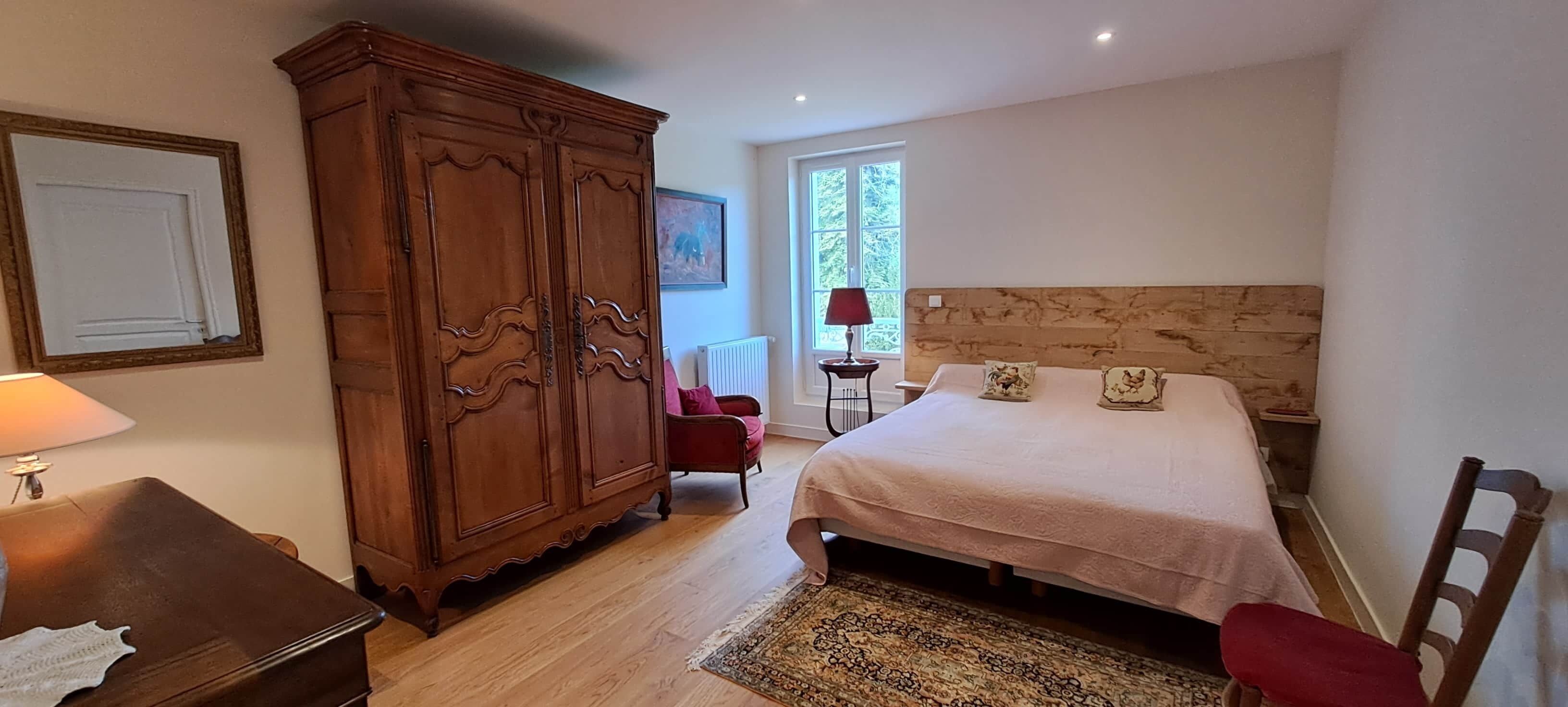 A bedroom with a large wooden wardrobe, a bed with a headboard, a red chair, and a window with natural light.