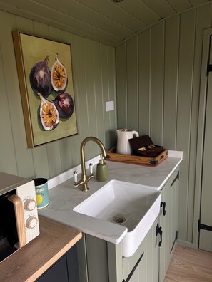 A kitchen with a white sink, brass faucet, and a painting of figs.