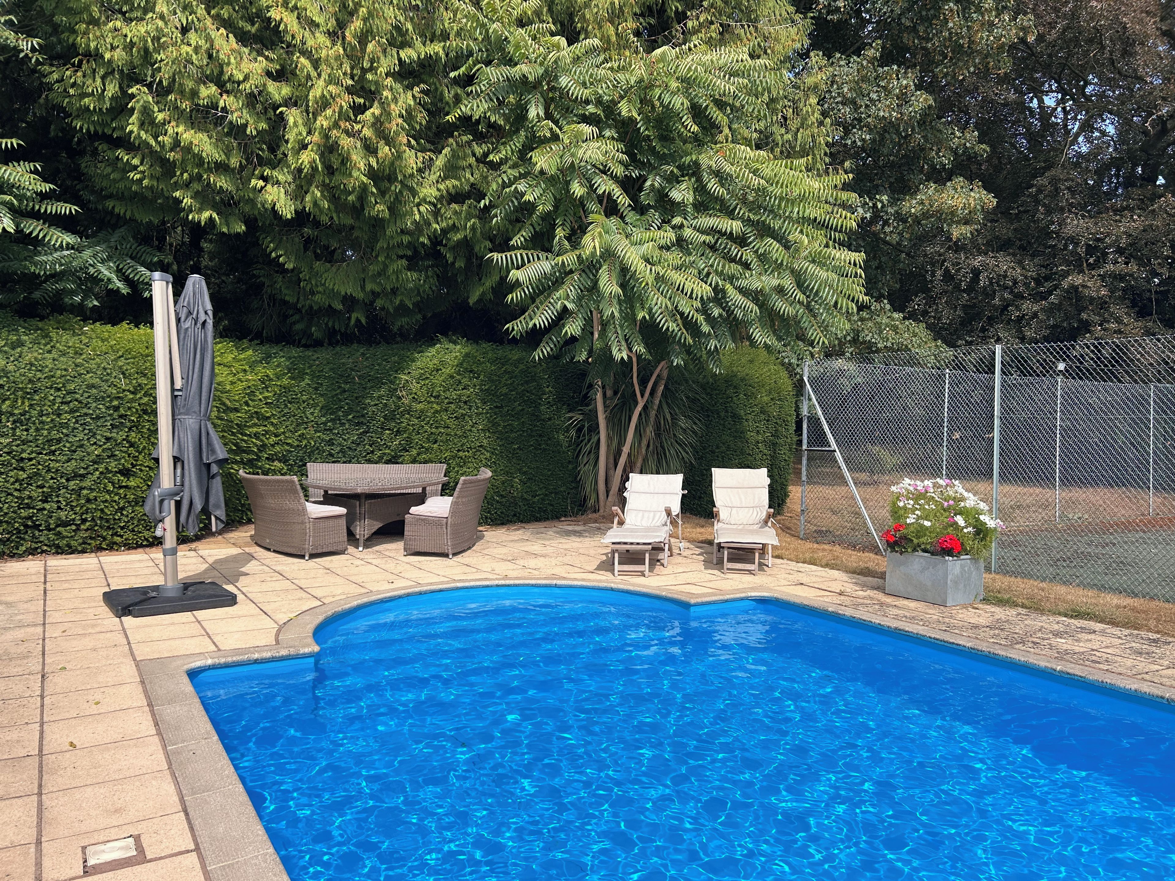 Une piscine entourée de verdure et d’une clôture, avec des chaises longues et une table à proximité.