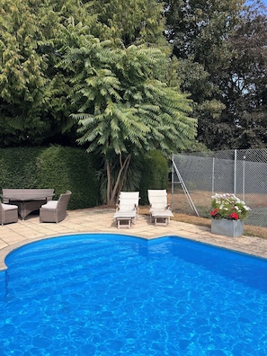 Une piscine entourée d’une haie et d’arbres, avec des chaises longues et une table à proximité.
