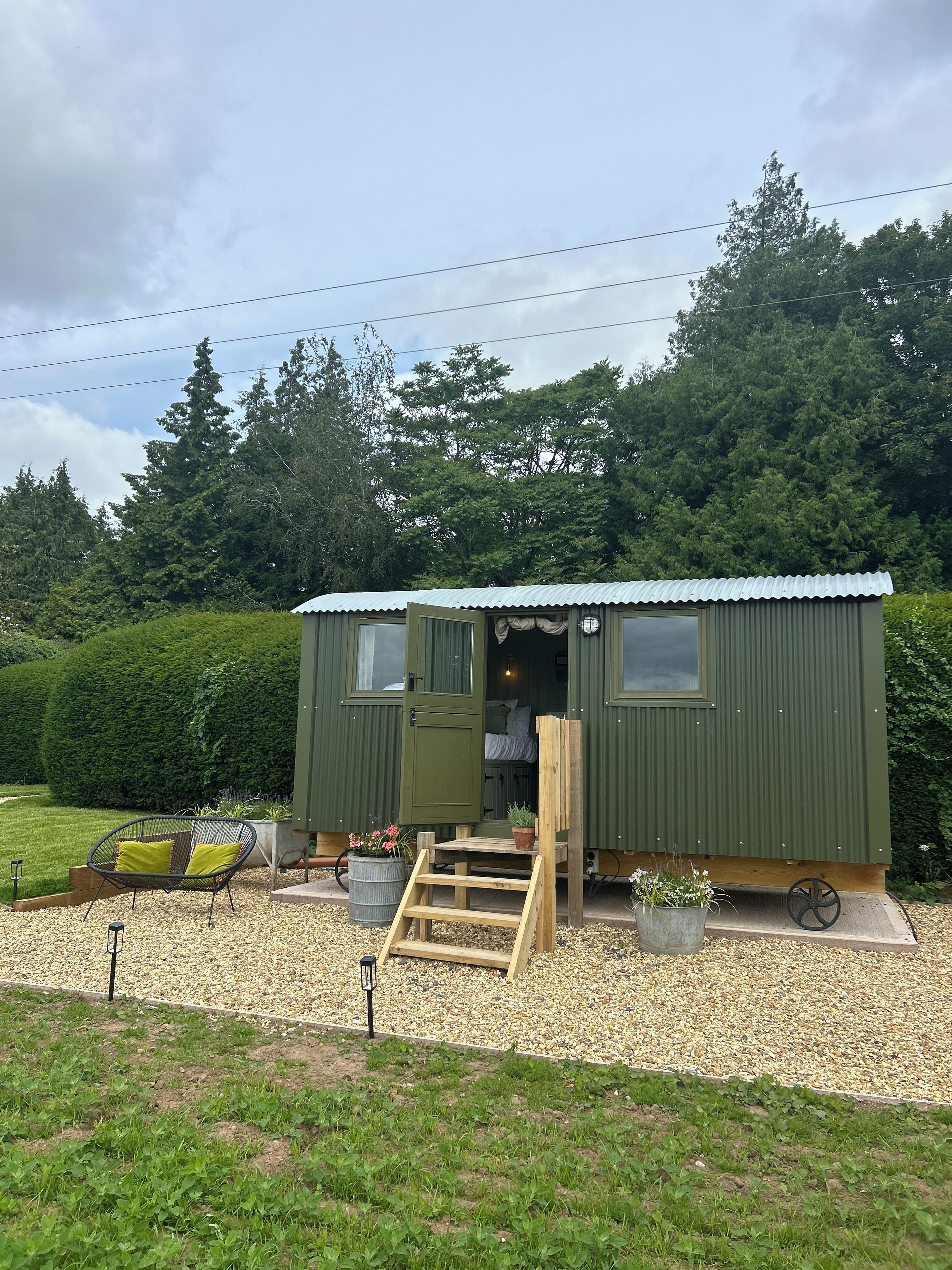 Une petite cabane à un seul étage, avec une façade verte, une porte en bois et un petit porche agrémenté d’un banc et de plantes en pot.