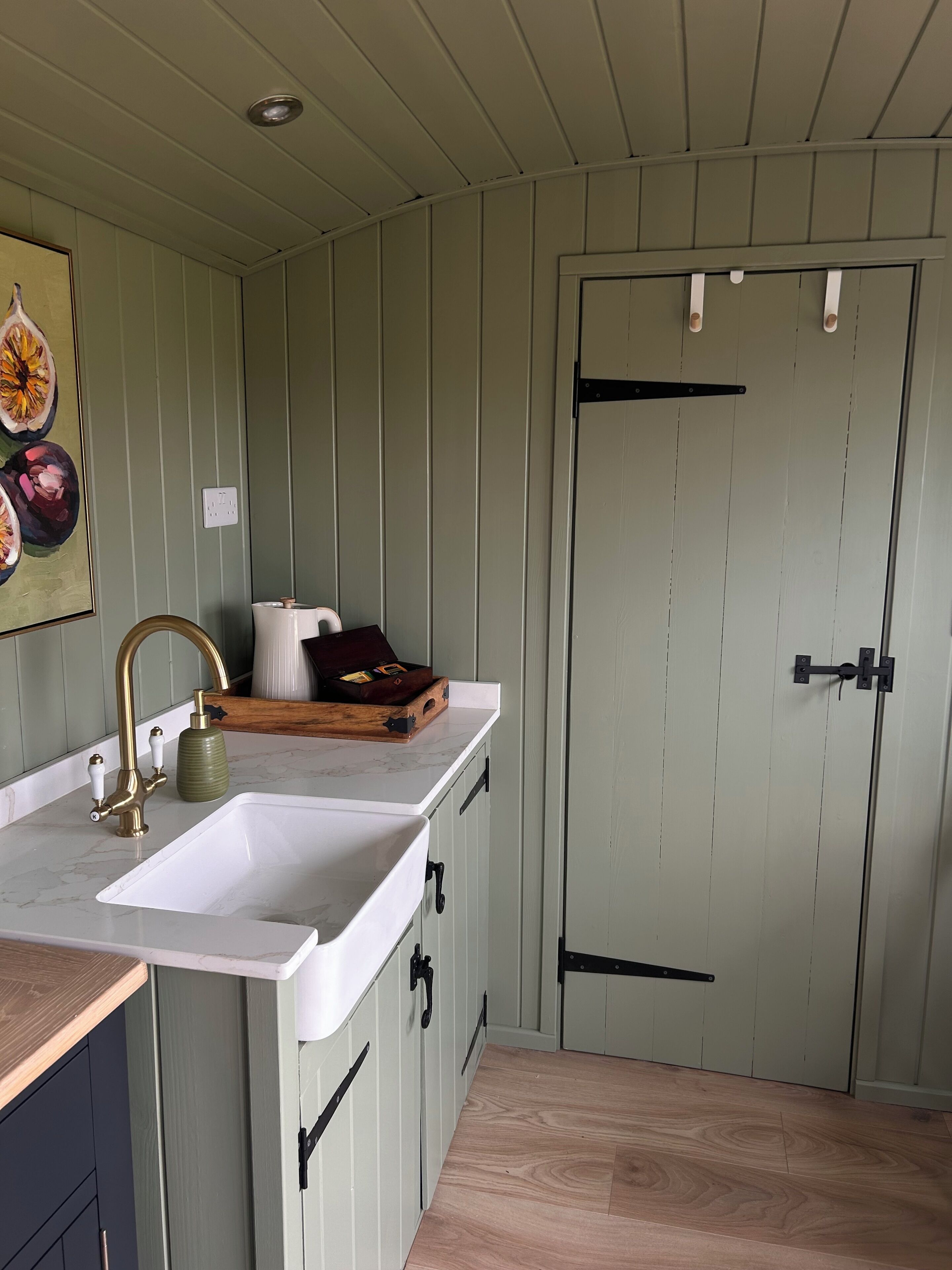 Une cuisine avec un évier blanc, un robinet en laiton et un plan de travail en bois. On y trouve une porte avec une poignée noire et un interrupteur blanc sur le mur.