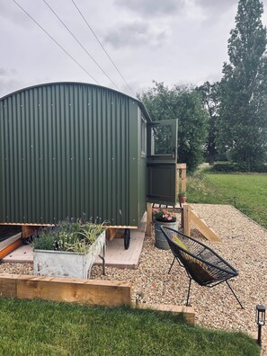 Un abri de jardin en métal vert avec une porte en bois, un petit banc en bois, un bac à plantes en béton avec des plantes, une chaise en métal noir et une zone recouverte de gravier.