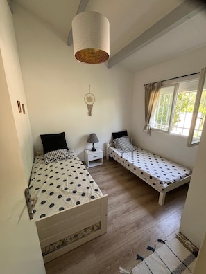 A bedroom with two single beds, a bedside table with a lamp, a hanging light fixture, a window with curtains, and a wall-mounted dreamcatcher. - Luxurious villa with a pool, close to the beaches and the harbor. (Saint-Cyprien)