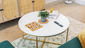 Ein runder weißer Tisch mit einem geometrischen Gesellschaftsspiel, einem kleinen Blumentopf, einer Fernbedienung und einem Buch, auf dem eine goldene Figur steht.