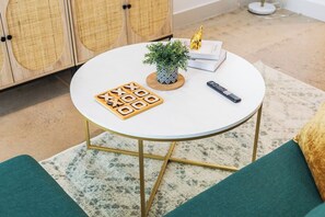 Ein runder weißer Tisch mit einem geometrischen Gesellschaftsspiel, einem kleinen Blumentopf, einer Fernbedienung und einem Buch, auf dem eine goldene Figur steht.