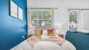 Ein ordentlich bezogenes Bett mit einem rosafarbenen Kopfteil, ein blaues Sofa und ein Fenster mit Jalousien.