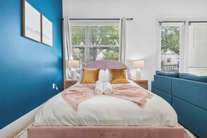 Ein ordentlich bezogenes Bett mit einem rosafarbenen Kopfteil, ein blaues Sofa und ein Fenster mit Jalousien.