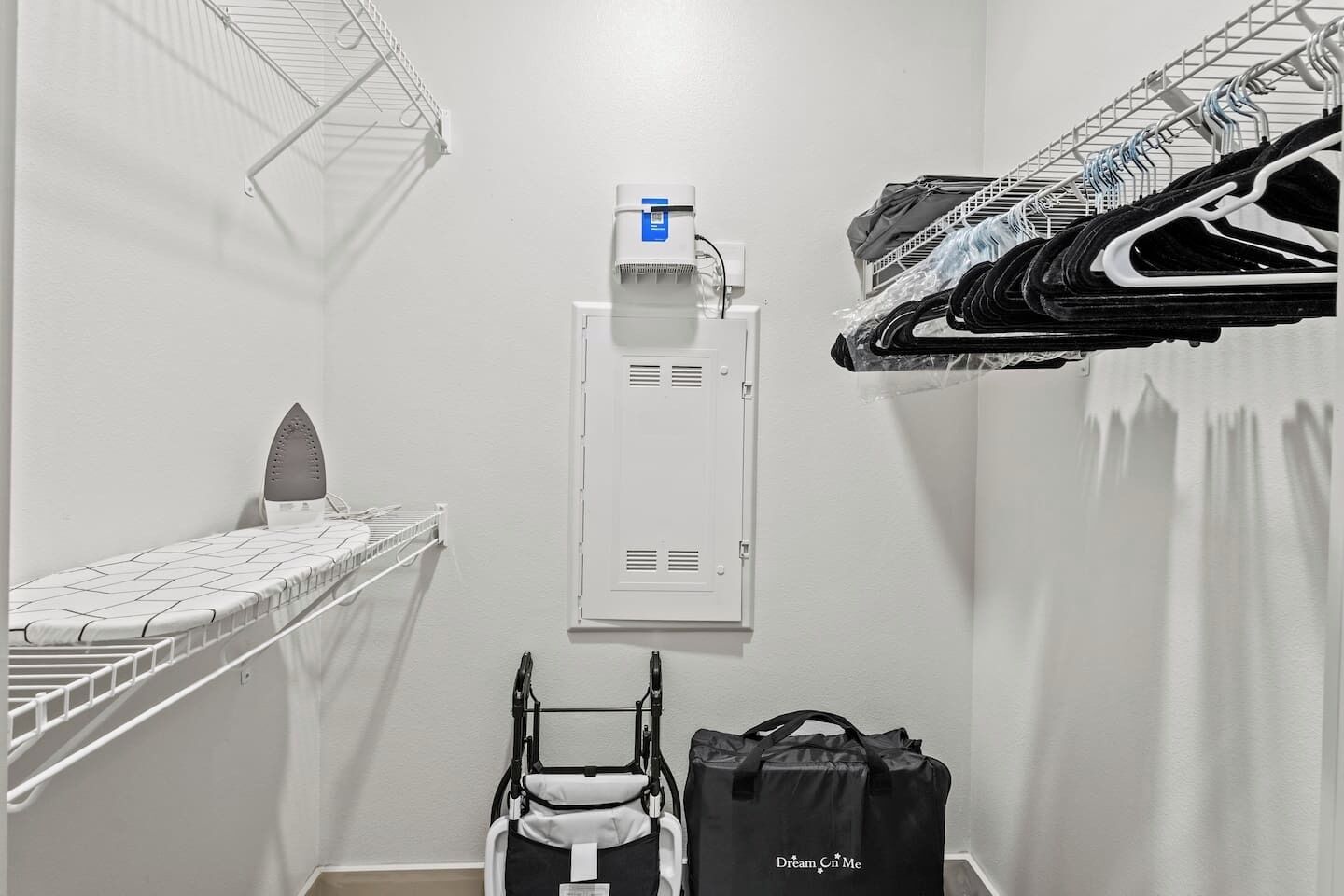 Une petite pièce avec un mur blanc, une petite planche à repasser, un radiateur fixé au mur, une poussette et un sac noir avec l’inscription « Dream on Me ».