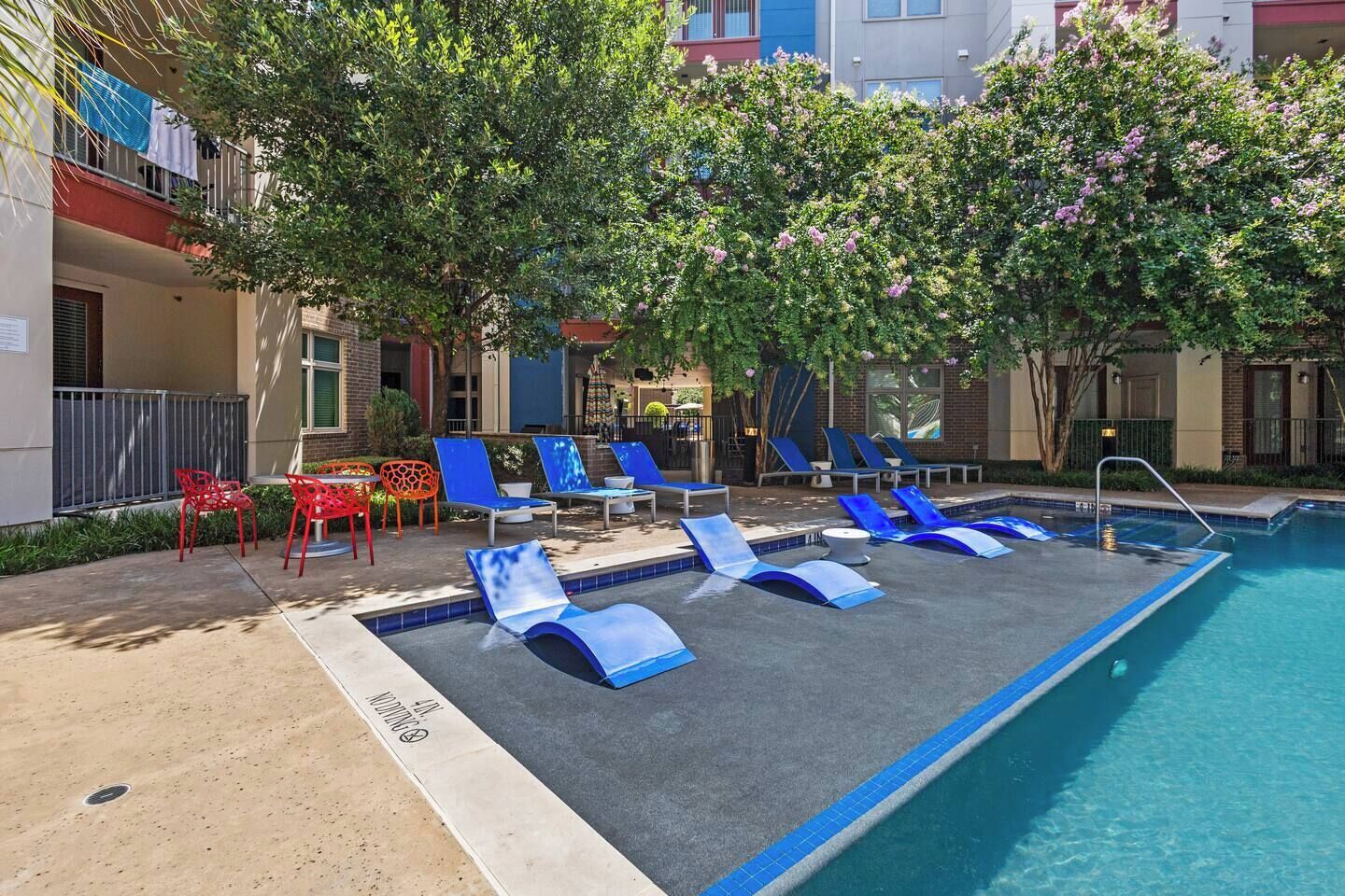 A pool area with lounge chairs and a small table, surrounded by trees and a building.