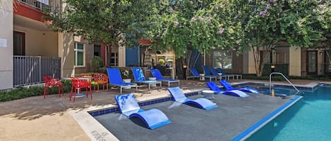 Area piscina con lettini e un piccolo tavolino, circondata da alberi e un edificio.