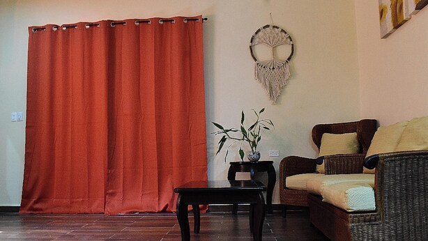 A room with a red curtain, a wall-mounted macrame piece, a small table with a plant, and a wicker sofa with cushions. - Home in tranquility and peace of the jungle situated in a beautiful garden. (Cozumel)