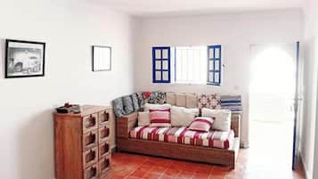 A room with a wooden chest of drawers, a sofa with patterned cushions, and a window with blue shutters.
