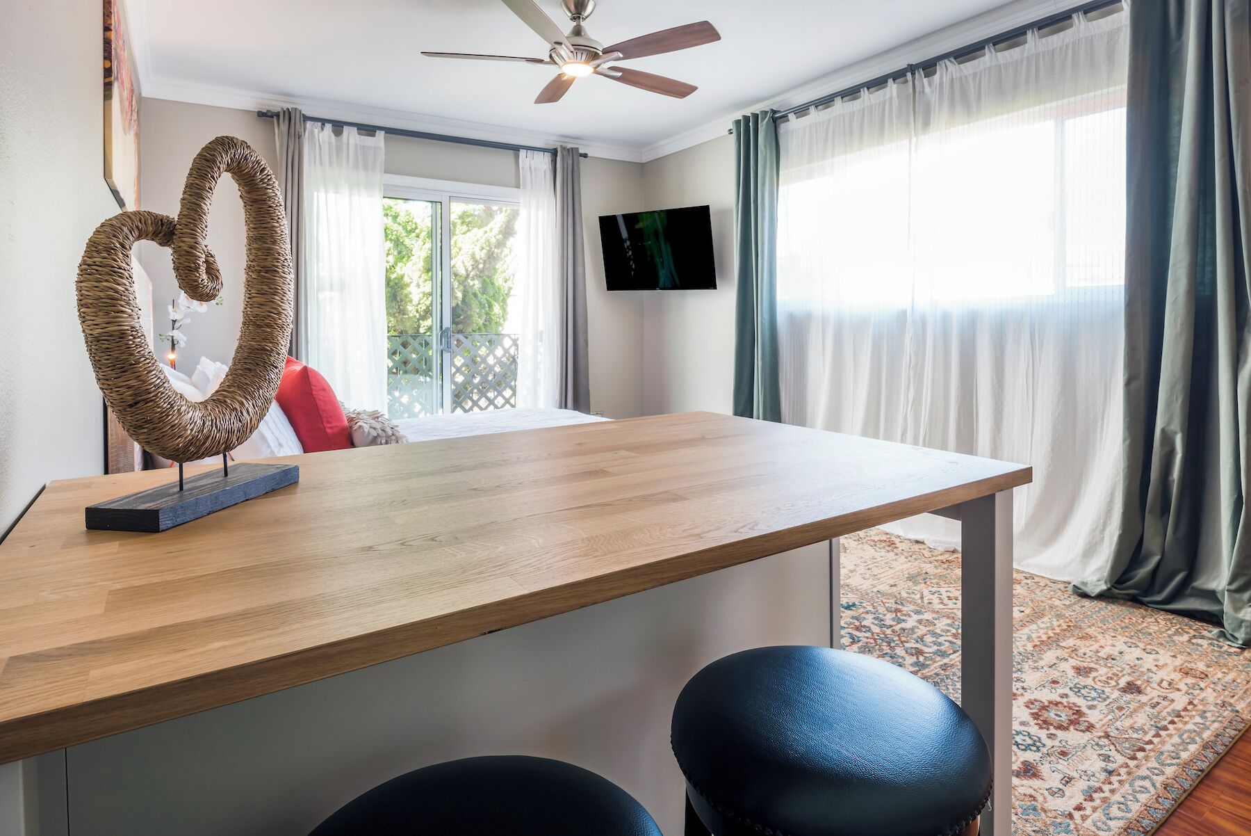 A modern room with a wooden table, two black leather chairs, a ceiling fan, a TV on the wall, and a view of greenery outside.