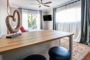 Ein modernes Zimmer mit einem Holztisch, zwei schwarzen Ledersesseln, einem Deckenventilator, einem an der Wand befestigten Fernseher und Blick ins Grüne.