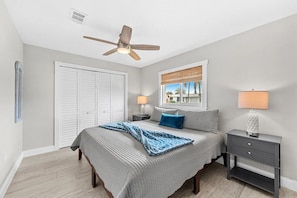A bedroom with a bed, bedside tables, a ceiling fan, a window with blinds, and a view of palm trees.