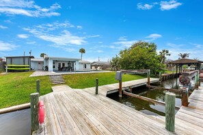 A wooden dock leading to a house with a pool, surrounded by a grassy yard and palm trees.