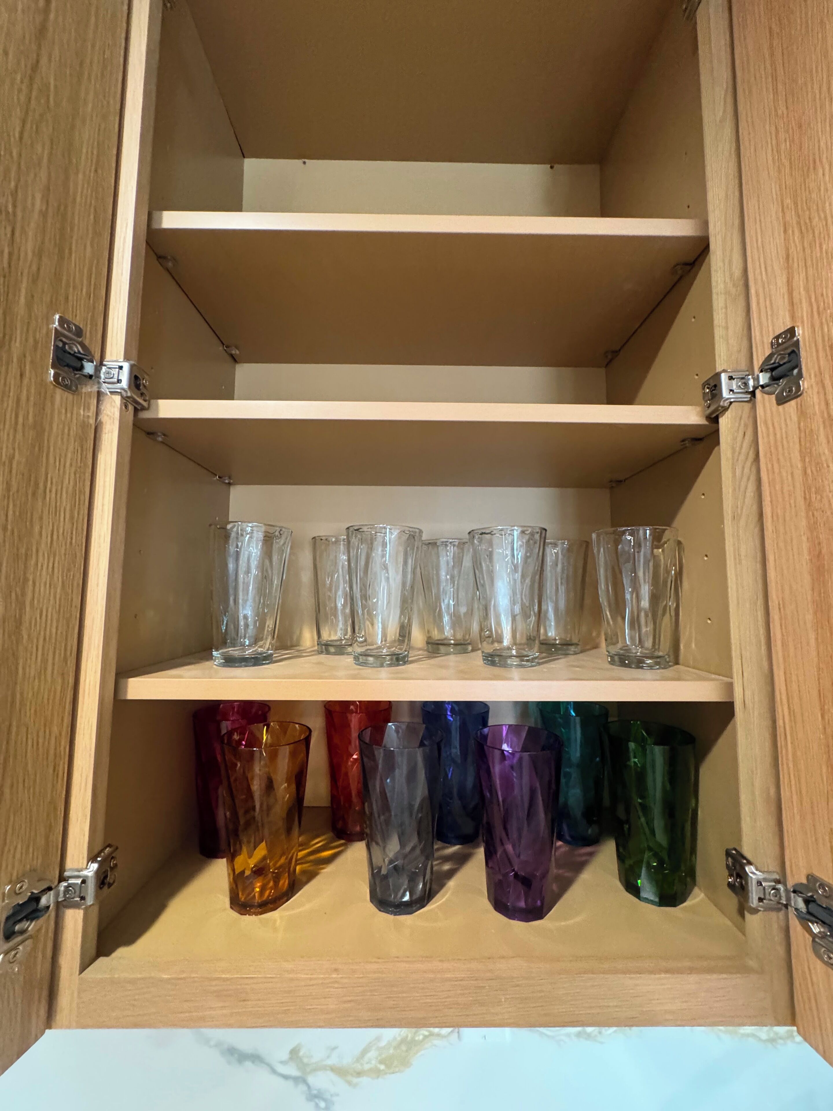 A wooden cabinet with two shelves; the top shelf holds clear glassware, and the bottom shelf contains colorful glassware.