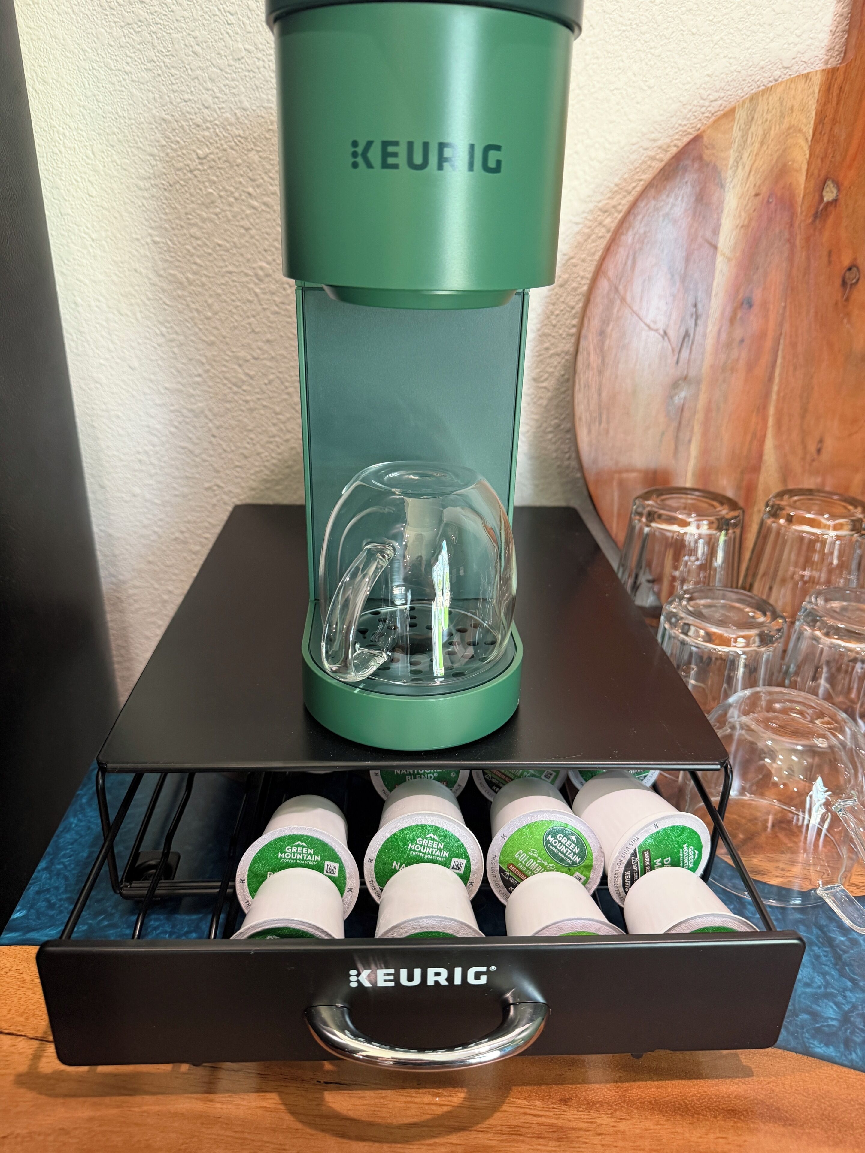 A Keurig coffee machine with a glass carafe on a black stand, a drawer with Keurig branded coffee pods, and several clear glass mugs in the background.