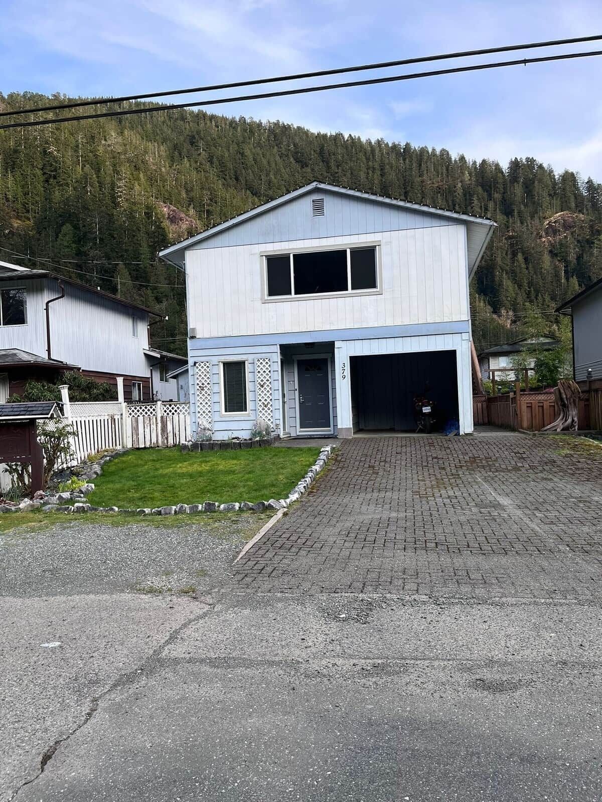 A two-story house with a garage, surrounded by a white picket fence, set against a forested hillside.
