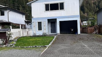 A two-story house with a garage, surrounded by a white picket fence, set against a forested hillside.