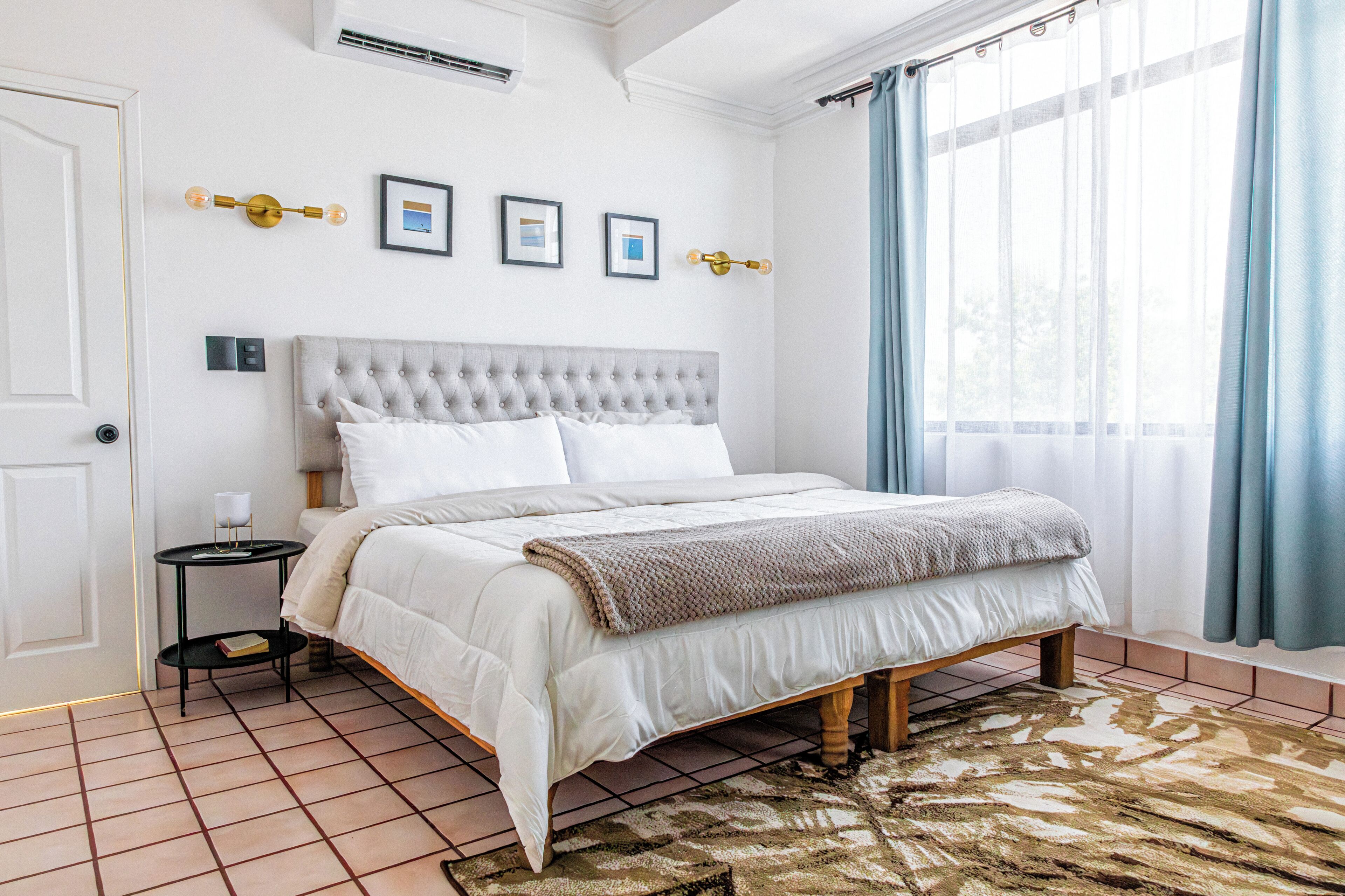 Un dormitori amb un panell de capçal amb borlons, un llit amb roba de llit blanca, una tauleta de nit amb una làmpada i una catifa amb dibuixos.