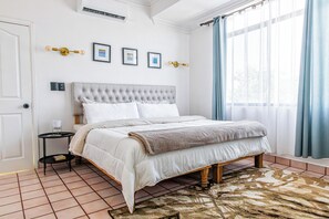 A bedroom with a tufted headboard, a bed with white bedding, a bedside table with a lamp, and a patterned rug. - Casa Palam 2 (La Paz)