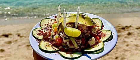 Una persona sosteniendo un plato con una ensalada en una playa, con agua azul cristalina al fondo.