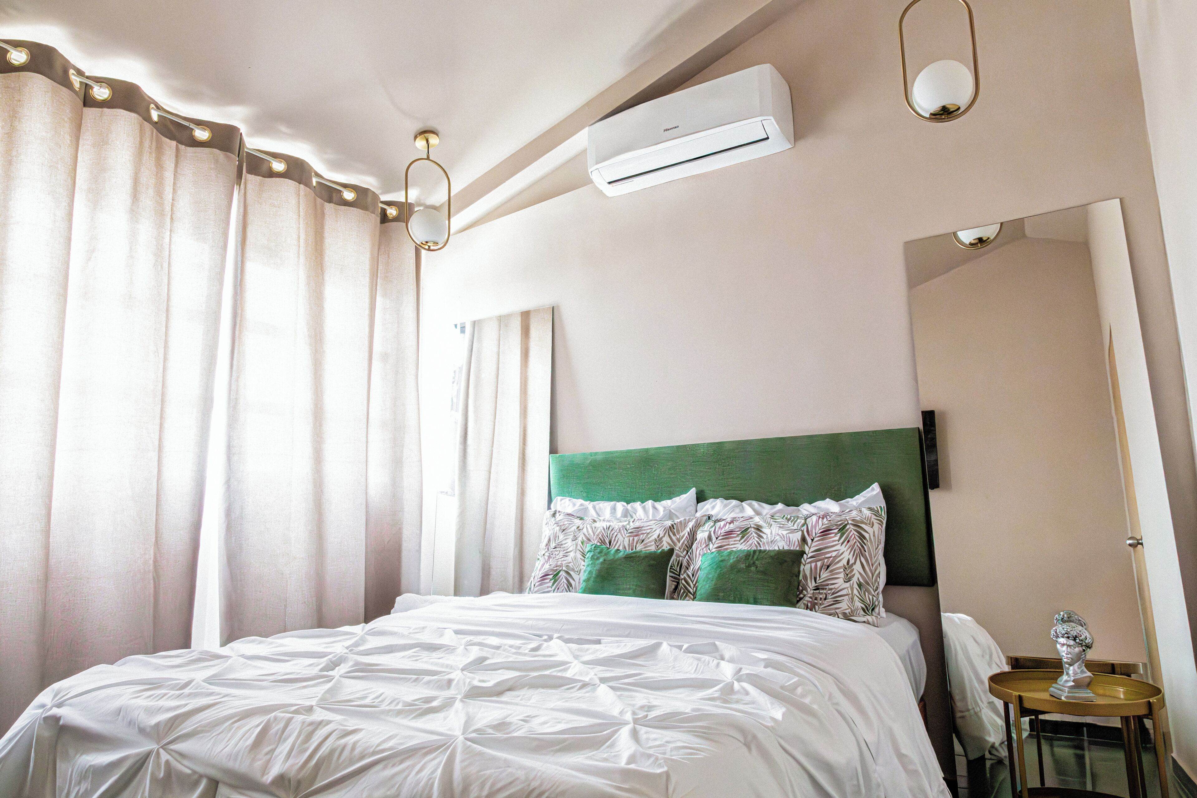 A neatly made bed with a green headboard, white bedding, and decorative pillows. An air conditioning unit is mounted on the wall above the bed.