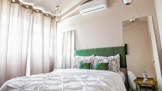 Un lit bien fait, avec une tête de lit verte, une literie blanche et des coussins décoratifs. Un appareil de climatisation est fixé au mur, au-dessus du lit.
