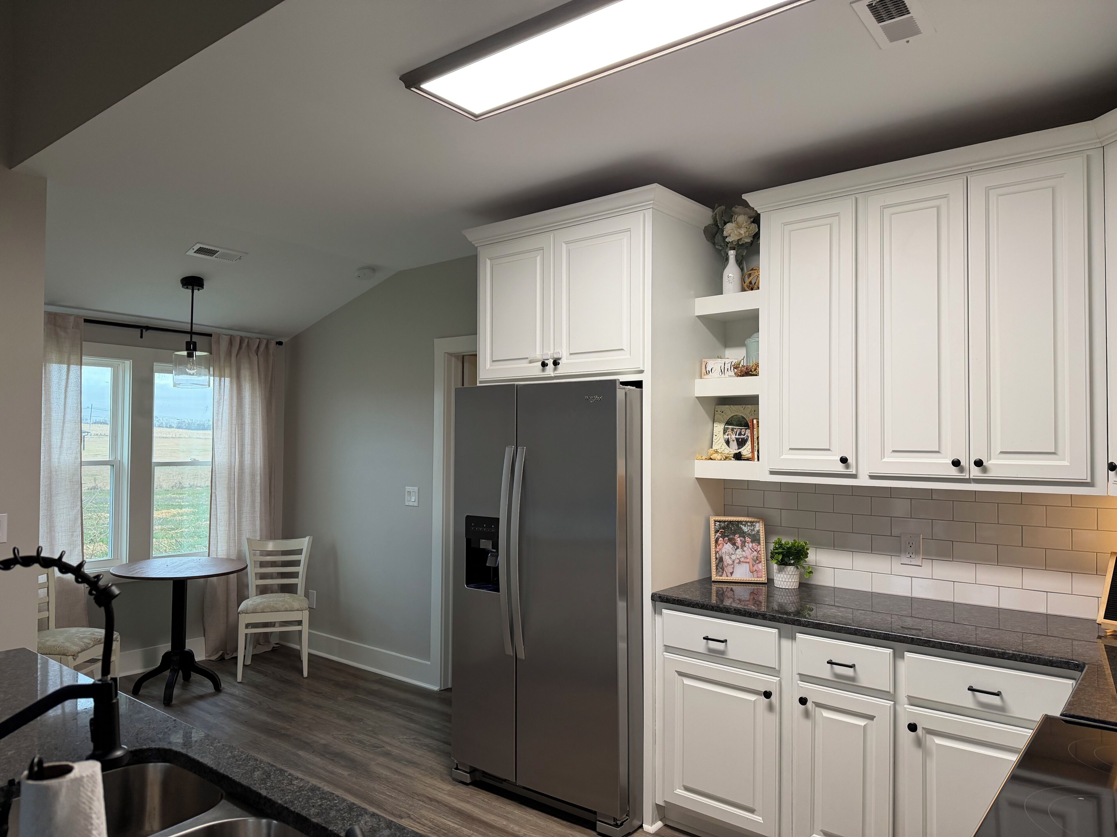 A modern kitchen with white cabinets, a stainless steel refrigerator, a dining area with a table and chairs, and a view through the window.