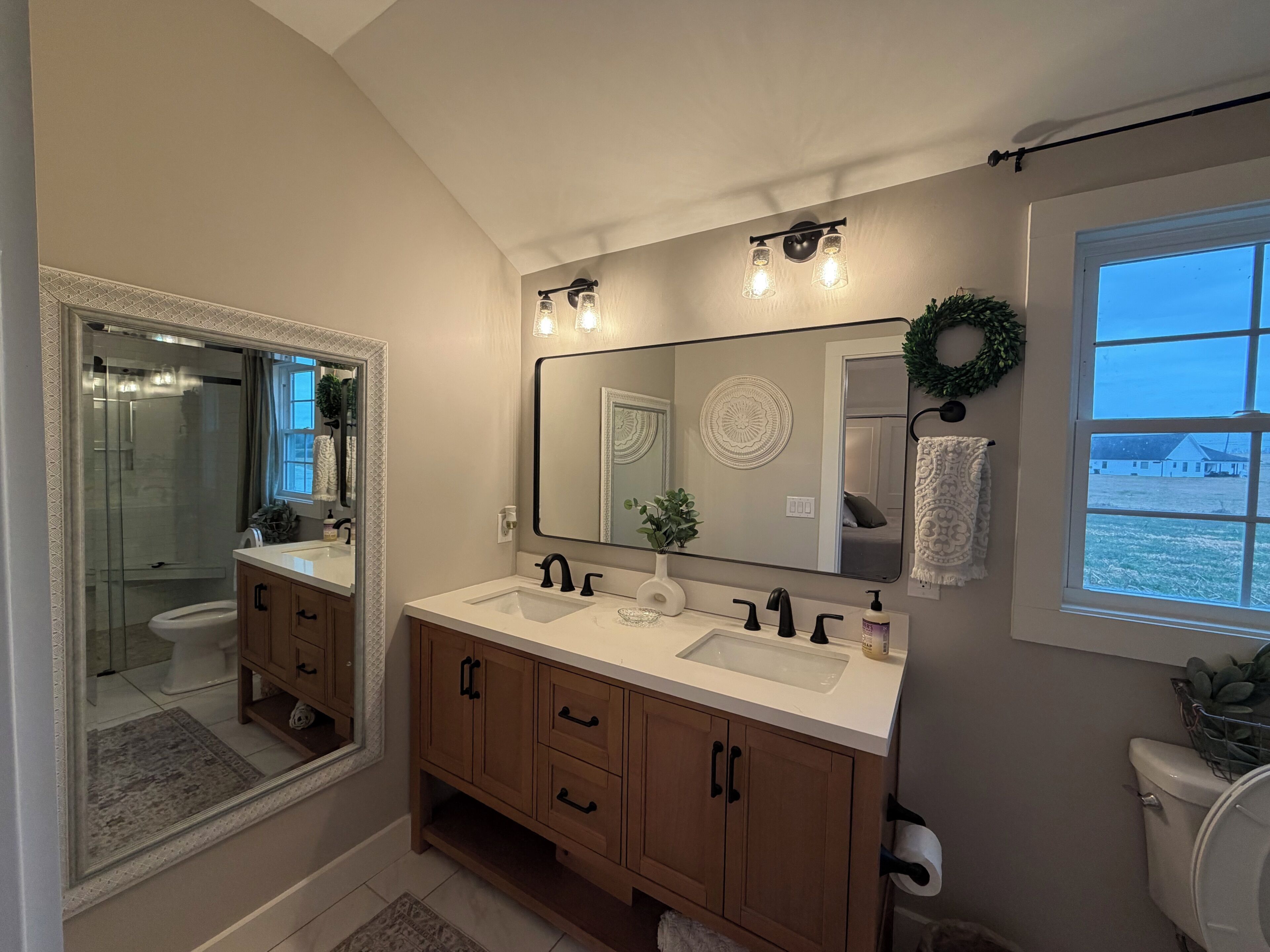 A bathroom with a double sink, a large mirror, a shower, and a window with a view.