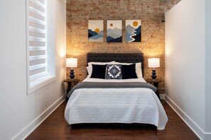 A bedroom with a brick wall, a bed with a quilt, two bedside lamps, and three wall art pieces.